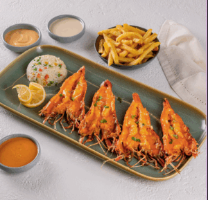 A plated seafood dish featuring four grilled prawns topped with a golden sauce, served with lemon slices, a portion of seasoned rice, French fries, and three small bowls of dipping sauces on a light-colored table.