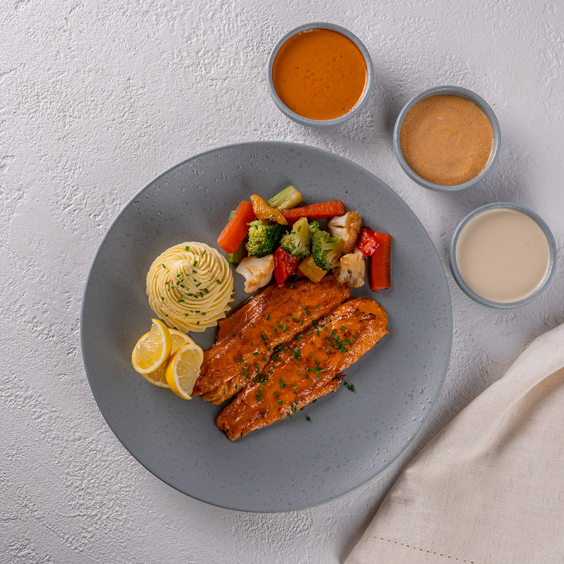 A plate of grilled salmon served with mashed potatoes, mixed sautéed vegetables, and lemon slices, accompanied by three small bowls of different sauces.