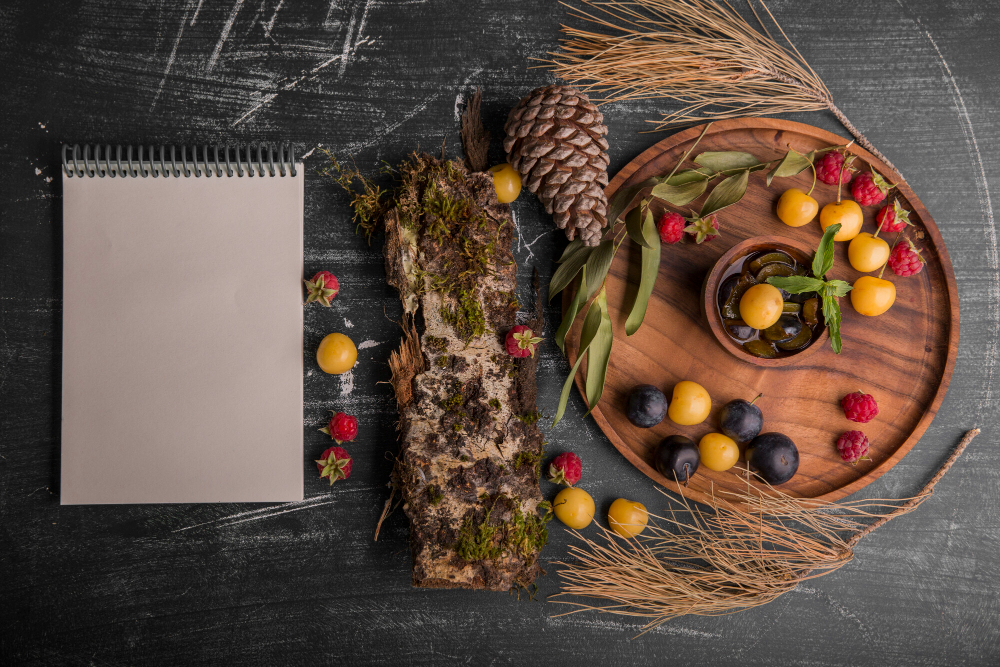A flat lay composition featuring a blank notepad, a rustic wooden log with moss, a round wooden tray filled with colorful fruits, and decorative natural elements like pinecones and dried wheat arranged on a dark surface.