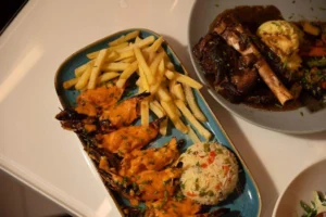 A platter of grilled prawns topped with a creamy orange sauce, served with French fries and a scoop of vegetable rice, with a second plate of braised lamb shank and mashed potatoes in the background.