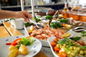 A person uses tongs to serve cooked shrimp from a buffet table filled with various seafood dishes, vegetables, and salads.
