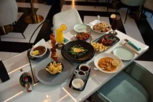 A beautifully set restaurant table displaying a variety of South African dishes at Villamoura Dubai. The spread includes flame-grilled skewered chicken served with fries, a creamy prawn curry, a plate of barbecued chicken wings, a fresh salad, sauces, and drinks garnished with lemon and orange slices. The elegant white and black tiled floor and modern gold-accented decor create a sophisticated dining atmosphere.